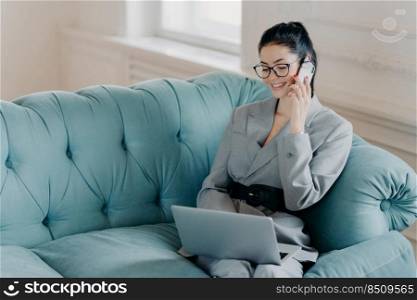 Pleased businesswoman in elegant clothes discusses financial issues with partner via mobile phone, poses on comfortable sofa, sits at modern laptop computer in home office. Technology, work concept