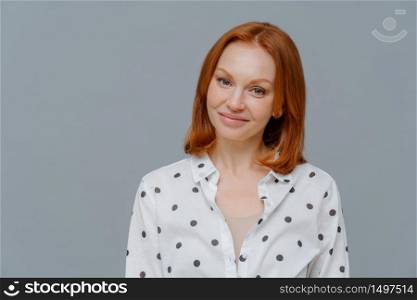 Pleased businesswoman with short red hair, makeup, looks positively at camera, has good mood after successful business meeting, dressed in stylish shirt, isolated over grey studio background.