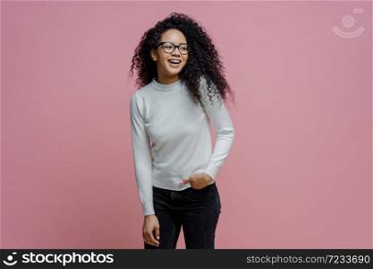 Pleased cheerful African American woman has happy carefree expression, keeps hand in pocket and looks aside with broad smile, wears white turtleneck and jeans, isolated over pink background.
