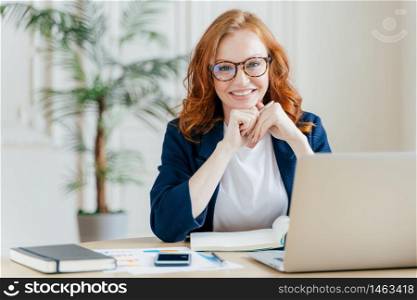 Pleased cheerful red haird female economist develops financial startup project, poses in office interior, works in business sphere, dressed in formal clothes, has happy expression, owns company