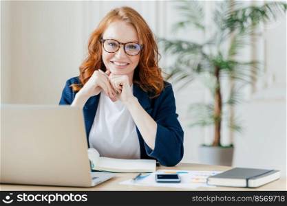 Pleased cheerful red haird female economist develops financial startup project, poses in office interior, works in business sphere, dressed in formal clothes, has happy expression, owns company