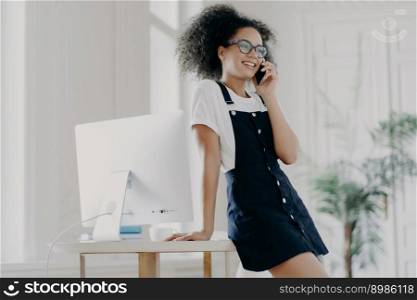 Pleased curly female office worker dressed in black sarafan, leans at table, poses in cabinet near computer monitor, has telephone conversation, discusses details of contract, has smile on face