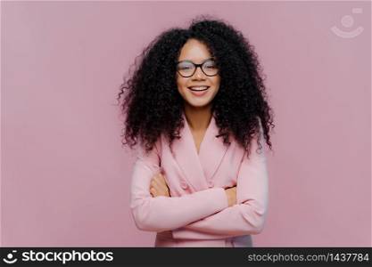 Pleased curly haired woman wears optical glasses for vision correction, elegant suit, keeps hands crossed over chest, poses against purple background, comes on job interview. Businesswoman at work