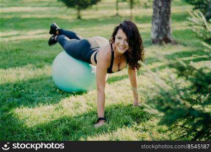 Pleased curly haired young woman with fit body leans on fit ball looks happily away dressed in sport clothes poses on green grass. Pretty European female does gymnastic exercises in open air