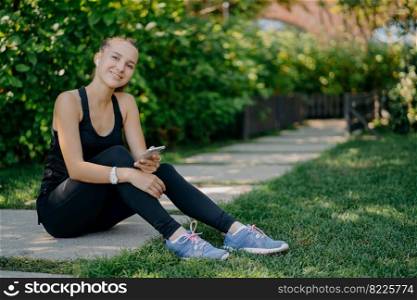 Pleased dark haired adult woman in sportsclothes uses smartphone for blogging and chatting online satisfied with workout results poses against nature background has break after fitness training