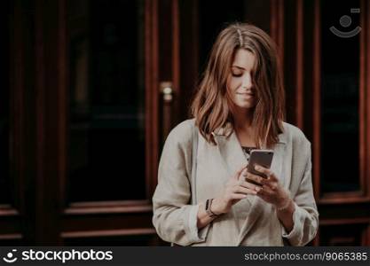 Pleased delighted female youngster watches video on smart phone, reads banking notification, dressed in white raincoat, poses against building interior. People, lifestyle and chatting concept