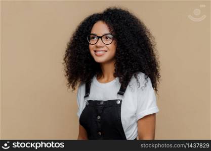 Pleased dreamy woman with curly hairstyle, looks through transparent glasses, dressed in casual white t shirt and black dungarees, poses against brown background, has good mood. People and emotions