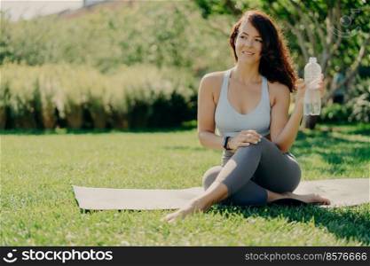 Pleased European woman poses on karemat and drinks water after workout stays in good physical shape looks somewhere spends free time doing sport outside. Summer time healthy lifestyle concept