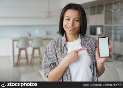 Pleased female customer showing phone with mock up empty screen looking at camera, standing at home. Smiling young woman pointing to smartphone with copy space, advertising mobile app, online services. Female customer shows mockup empty phone screen, advertises apps, online service, standing at home