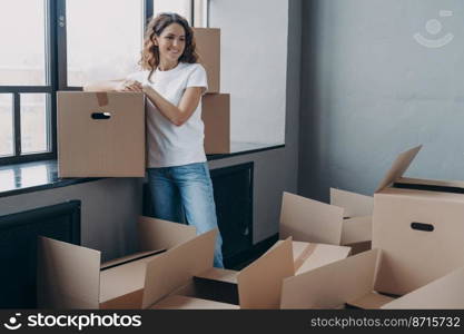 Pleased female homeowner standing with cardboard boxes in empty room. Spanish woman with many cartons for things on day of relocating to new home. Relocation, moving and delivery concept.. Female homeowner standing with cardboard boxes on day of relocation to new home. Moving and delivery