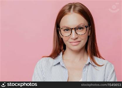Pleased female journalist wears elegant shirt, optical glasses, has full lips, pleased to meet with people, poses against pink studio wall with blank space for your promotional content. Indoor shot
