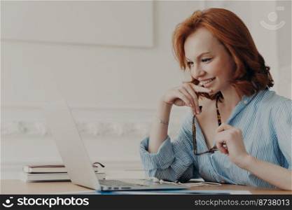 Pleased female model poses at coworking space, holds spectacles, smiles positively, uses wireless internet connection, reads information, has online communication, works on creating web project