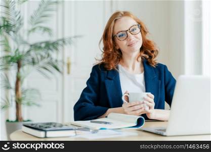 Pleased female teacher makes research work on laptop computer, writes down information in notepad, enjoys drinking coffee, has happy smile on face, sits in spacious room at desktop plant in background