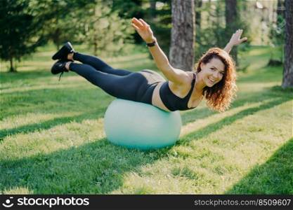 Pleased fit woman excercises outdoor on fitness ball raises arms tries to balance dressed in active wear has perfect healthy body does yoga exercises outdoor on green grass. Healthy sporty lifestyle