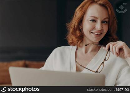 Pleased ginger female student spends time on online education, uses laptop computer and wireless connection, concentrated on remote job, holds spectacles, smiles toothily, installs new application