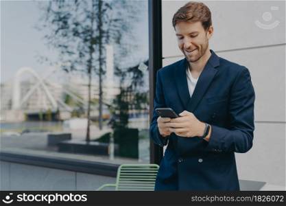 Pleased handsome male CEO or director holds mobile phone waits for call scrolls social networks while waiting for partner near outdoor cafe wastes time online dressed in elegant formal suit.. Pleased handsome male CEO or director holds mobile phone waits for call