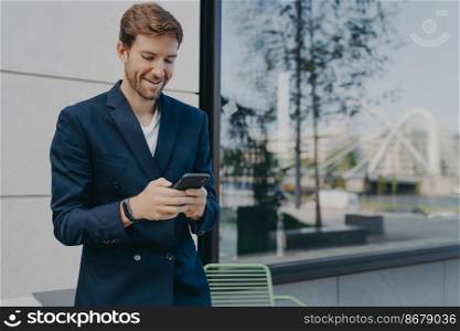 Pleased handsome male CEO or director holds mobile phone waits for call scrolls social networks while waiting for partner near outdoor cafe wastes time online dressed in elegant formal suit.. Pleased handsome male CEO or director holds mobile phone waits for call