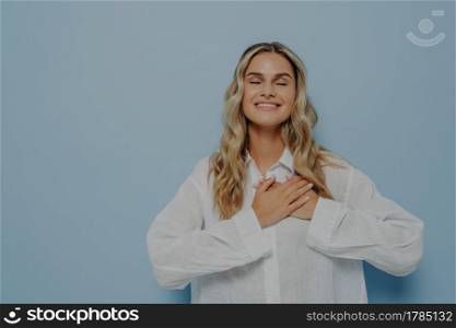 Pleased happy thankful woman with blond wavy hair in white oversize shirt gesturing appreciation and love and gratitude by holding hands together on chest, eyes closed, isolated over blue wall. Happy thankful woman with blond wavy hair in white oversize shirt gesturing appreciation and love