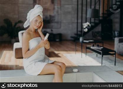 Pleased healthy woman applies body lotion and face cream, nourishes skin, wrapped in bath towel, smiles gently, poses against domestic interior, undergoes beauty treatments. Dermatology concept