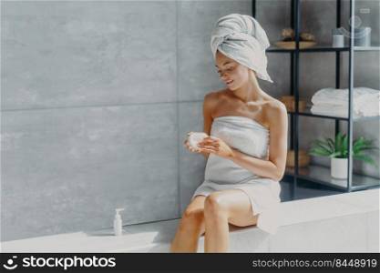 Pleased healthy woman wrapped in bath towel applies body cream after showering takes care of skin poses in bathroom undergoes beauty treatments has hygiene routine. P&ering and wellness concept