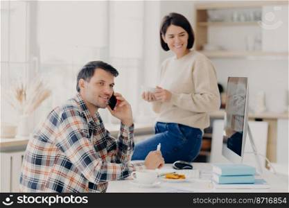 Pleased husband and wife manage finances at home, review bank accounts, pleased man holds mobile phone near ear, wears plait shirt, pleased woman sits on table, drinks aromatic coffee from mug