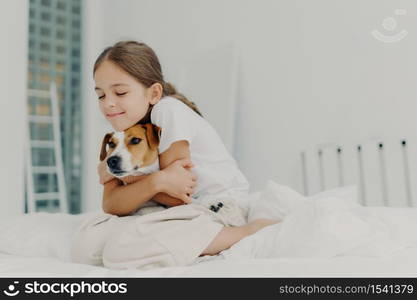 Pleased little girl plays with pet, embraces dog and keeps eyes closed from pleasure, dressed in nightclothes, poses on bed after waking up, expresses love to pedigree animal. Bedding concept