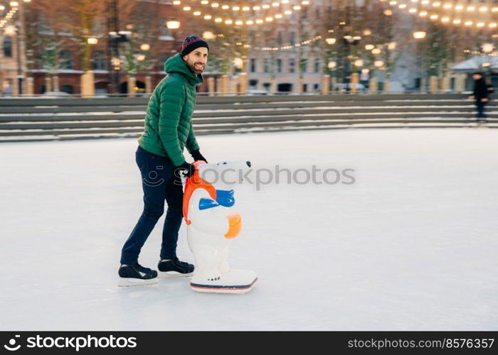 Pleased male skater being on skating ring, uses skate aid as tries to be in balance, looks happily aside, poses on ice. Cheerful man dressed in warm clothes, involved in active lifestyle.