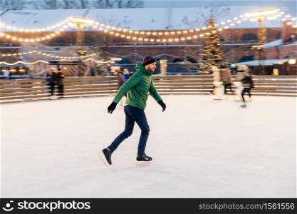 Pleased man in green jacket, wears skates, goes skating on ice, spends winter holidays with use, demonstrates his professionalism, enjoys snowy weather. Winter fun outdoor activities