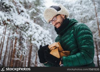 Pleased man in spectacles looks hapily at firewood which he log in winter forest, going to make fire, wears green anorak, poses against white winter forest. People, lifestyle, recreation concept