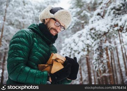 Pleased man in spectacles looks hapily at firewood which he log in winter forest, going to make fire, wears green anorak, poses against white winter forest. People, lifestyle, recreation concept