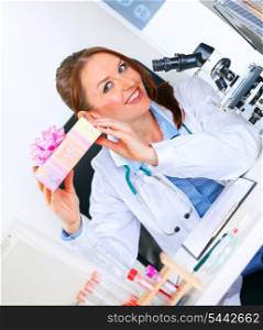 Pleased medical doctor woman sitting at office table and holding gift in hands&#xA;