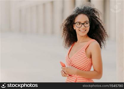 Pleased millennial girl with curly hair, wears pink dress, optical glasses, focused aside with smile, stands outdoor, waits for call, enjoys high speed internet connection, updates new application