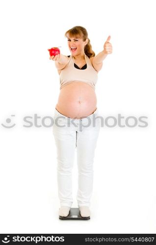 Pleased pregnant woman standing on weight scale and showing thumbs up isolated on white&#xA;