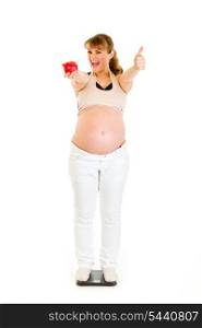 Pleased pregnant woman standing on weight scale and showing thumbs up isolated on white&#xA;