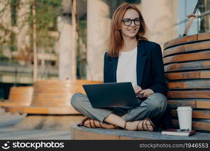 Pleased pretty redhead woman learns educational course on laptop computer sits crossed legs on wooden bench outdoors drinks takeaway coffee looks happily into distance creats new publication. Pleased pretty redhead woman learns educational course on laptop computer