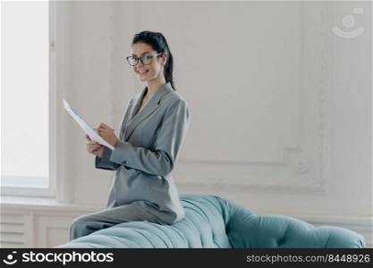 Pleased prosperous woman financial manager studies documents in spacious home office, leans at sofa, dressed in elegant clothes, looks gladfully at camera, writes information. Paper work concept