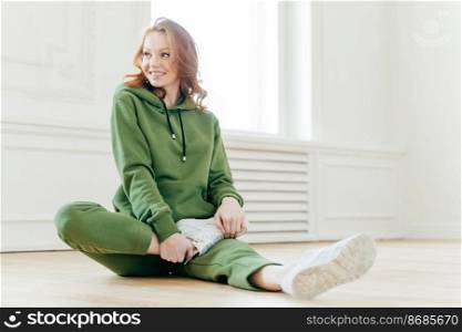 Pleased redhead woman focused aside, has happy expression, stretches legs, sits on floor, waits for instructor, wears hoody, white sneakers, has yoga exercising. People and motivation concept