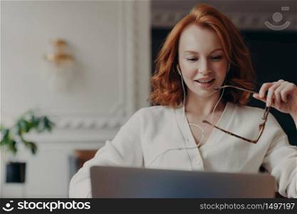 Pleased redhead woman student watches training webinar, has online video conference, uses earphones, works on business project, keeps transparent glasses in hand. Distance work via internet.