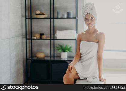Pleased relaxed smiling woman with bath towel around healthy naked body, poses in bathroom with cosmetic product, takes care of herself, has skin care treatments. Spa, wellness, hygiene concept