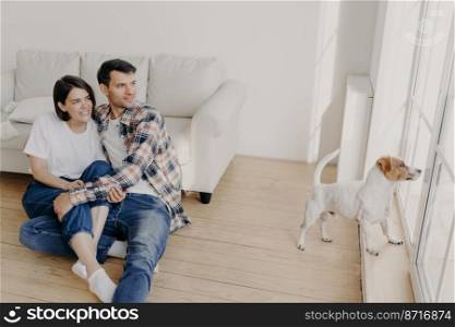 Pleased satisfied romantic couple embrace and looks out of window, pose in empty room near sofa, their dog wants to have outdoor walk. Woman and man enjoy spare time at home, live in new flat