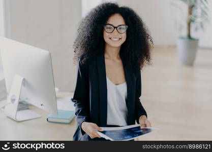 Pleased skilled dark skinned curly woman holds some papers, poses in modern office interior, wears transparent glasses and formal suit, stands near table with computer, studies documentation