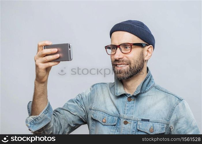 Pleased smiling bearded male with happy expression poses at camera of smart phone, takes photo of himself, wears denim jacket and glasses, isolated over white background. People and technology concept