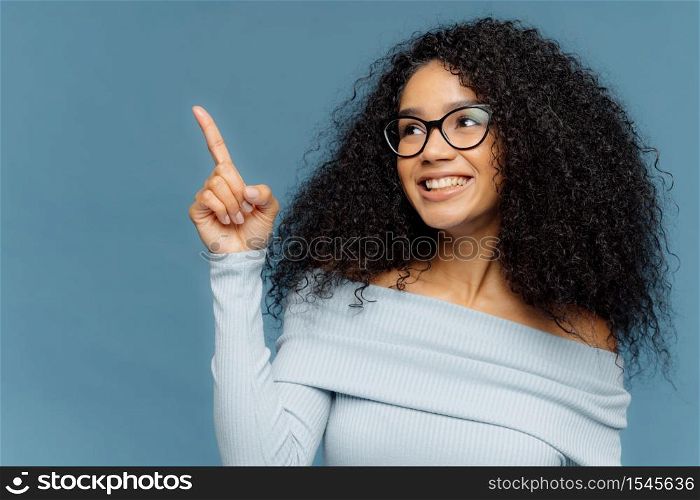 Pleased smiling dark skinned female with Afro hairstyle points index finger upwards, demonstrates something on blank space, has glad expression, wears blue sweater, stands indoor. People and promotion