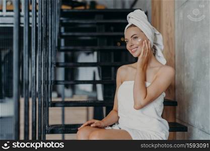 Pleased smiling woman applies face cream, poses on stairs, wrapped in bath towel, smiles gently and has minimal makeup, enjoys softness after taking shower, takes care of skin. Wellness concept