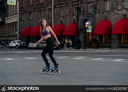 Pleased sporty woman rides on rollers in urban place leads healthy lifestyle dressed in sportsclothes looks away has cheerful expression enjoys fitness training. Sports activity hobby recreation