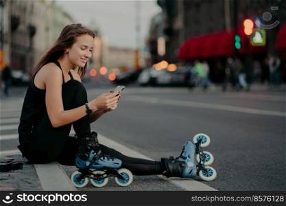 Pleased sporty woman wears sportsclothes rollerblades sits on road checks newsfeed via smartphone takes break after inline skating poses against blurred city background engaged in healthy lifestyle