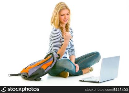 Pleased teengirl sitting on floor with schoolbag and laptop isolated on white &#xA;