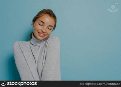 Pleased tender woman keeps eyes closed smiles tenderly recalls nice moment from life dressed in casual turtleneck stands against blue background with copy space for your promotional content.. Pleased tender woman keeps eyes closed smiles tenderly recalls nice moment from life