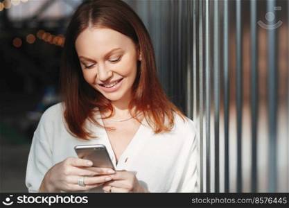 Pleased woman focused into screen of cell phone, checks email box, dressed in white clothes, sends feedback, connected to wireless internet, has brown hair, charming smile, sends text message