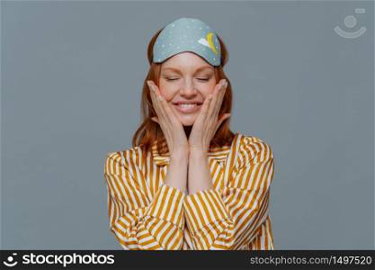 Pleased woman touches cheeks, has healthy freckled skin, smiles broadly prepares for sleep, wears striped pajama and sleep mask, closes eyes, isolated over grey background. Rest and lifestyle concept